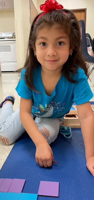 A young girl with long dark hair and a red bow sits on a blue yoga mat, surrounded by colorful paper shapes on the floor.