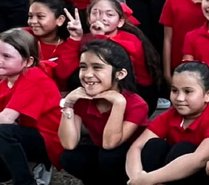 A group of young children wearing red shirts are gathered together, smiling and making gestures, against a dark background.