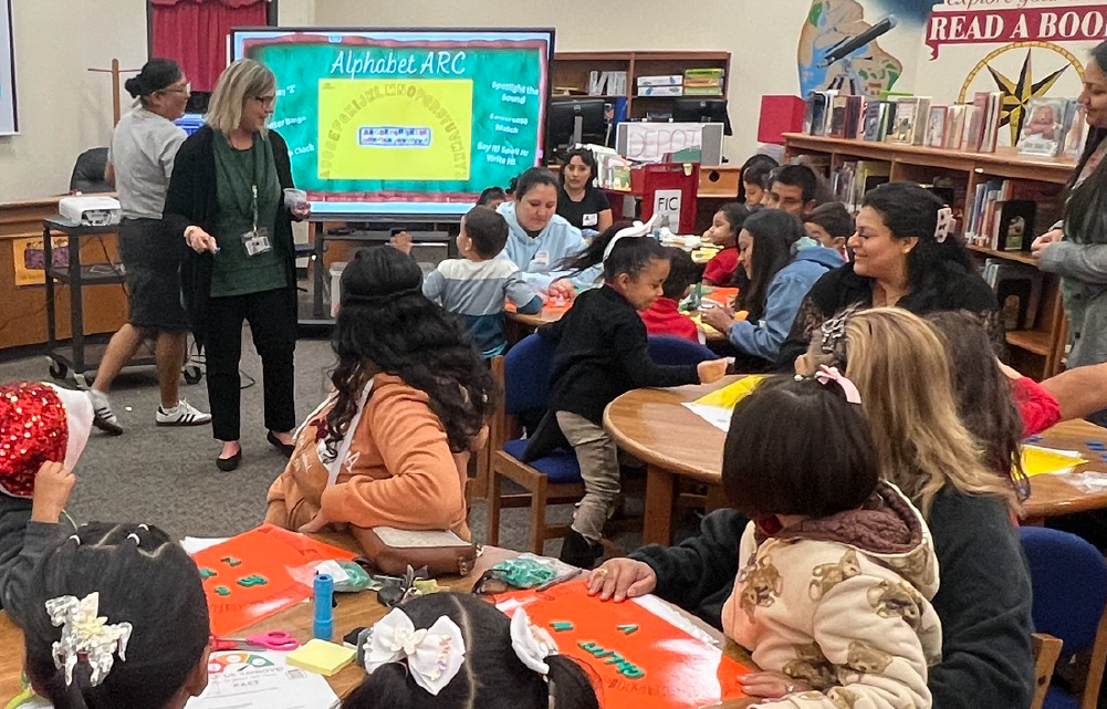 A group of students and a teacher are gathered around a table in a classroom, engaged in what appears to be a collaborative learning activity or discussion.