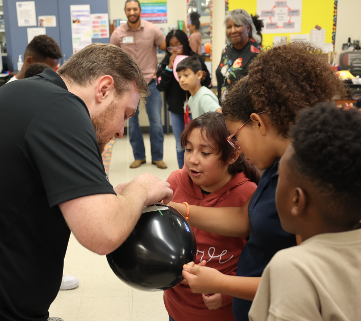 A group of children, some wearing red shirts, are gathered around an adult man who is helping them put on boxing gloves in what appears to be a classroom or community center setting.