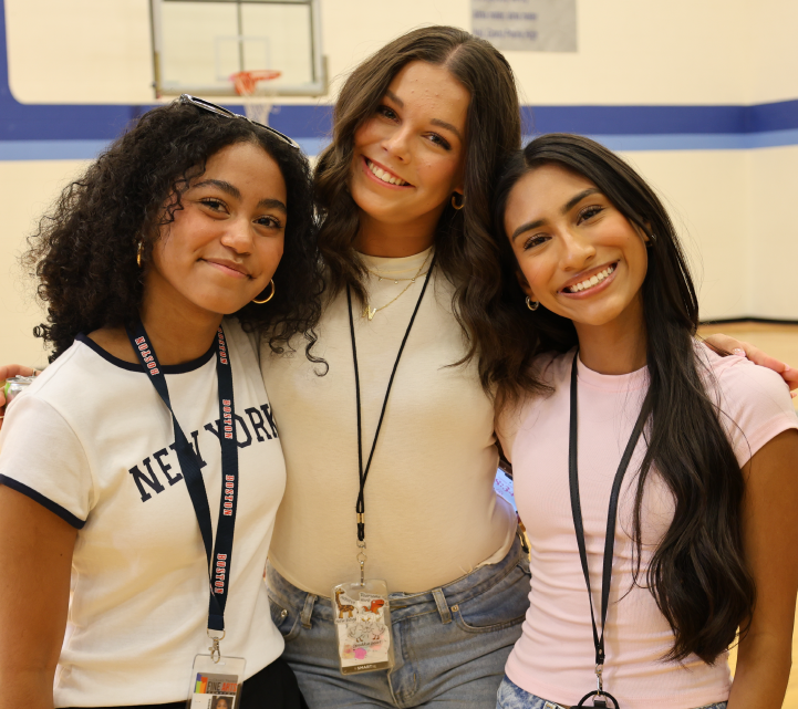 Three young women with diverse appearances are smiling and posing together in what appears to be a school or educational setting, with a blue wall and signage visible in the background.