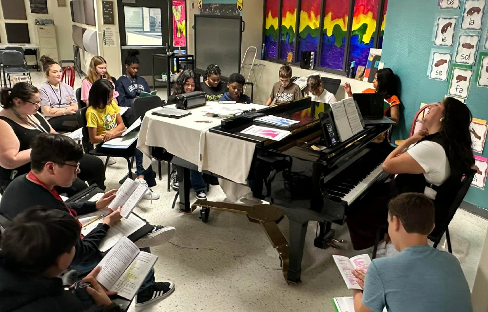 A group of people, likely students, gathered around a piano in a classroom setting, with colorful artwork and posters adorning the walls in the background.