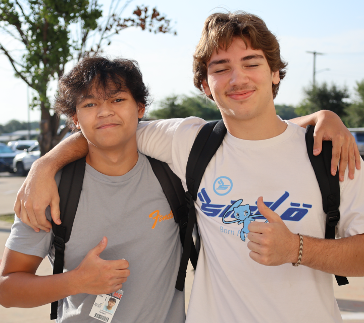 Two young men, one wearing a gray shirt and the other a white shirt with a logo, are standing together and smiling in an outdoor setting with trees and a parking lot visible in the background.