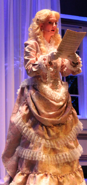 A woman in an elaborate, ornate dress stands on a stage, holding a document in her hands and appearing to be in the midst of a performance or presentation.