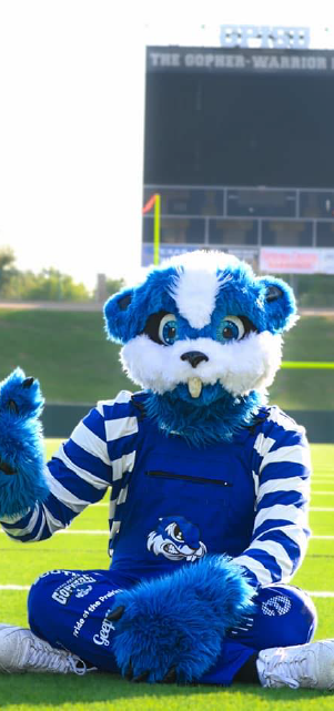 A blue and white furry mascot character stands on a sports field with a scoreboard visible in the background.