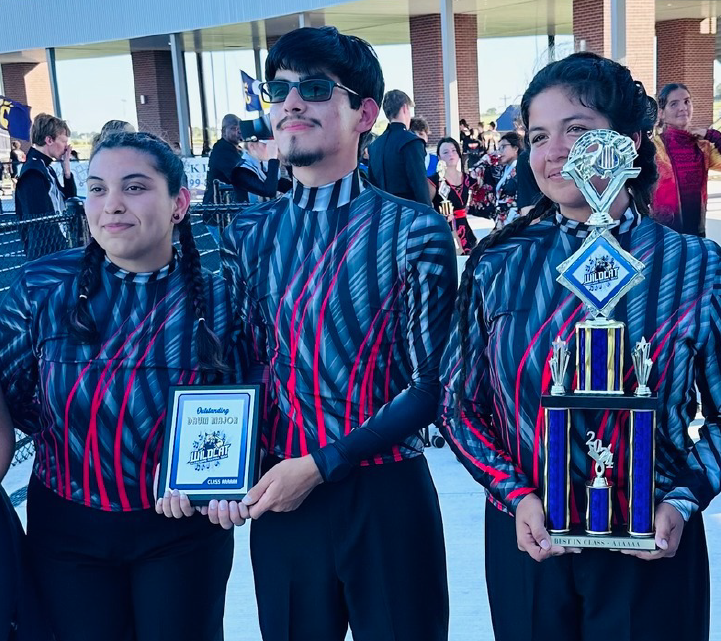 The image shows three individuals, two males and one female, holding trophies and awards, standing in front of a crowd of people. They appear to be celebrating some kind of achievement or victory.