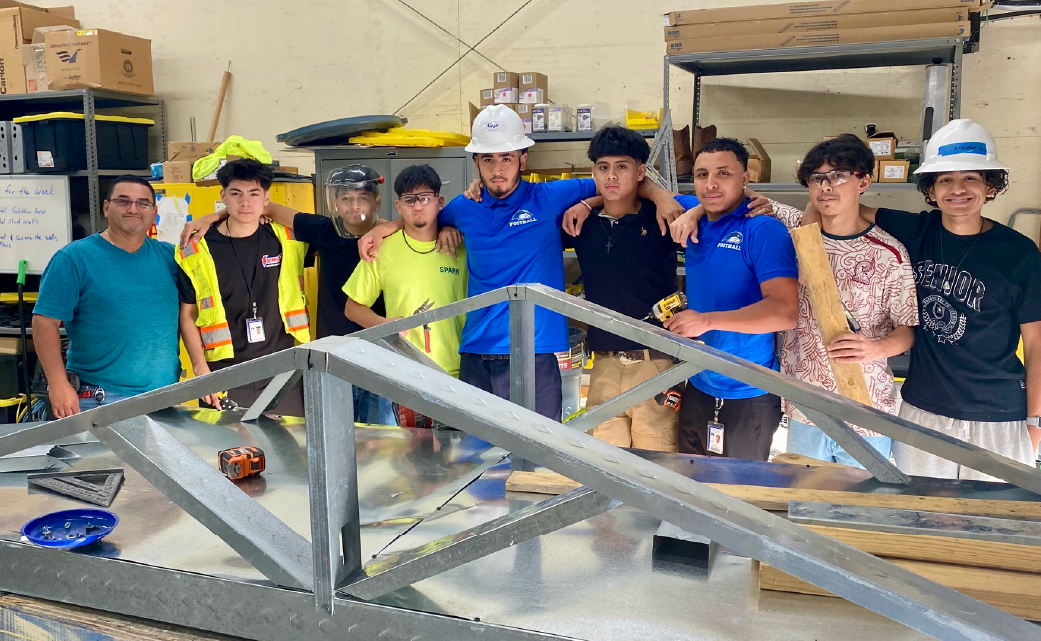 A group of people, some wearing safety gear, standing around a metal structure in what appears to be a workshop or construction site.