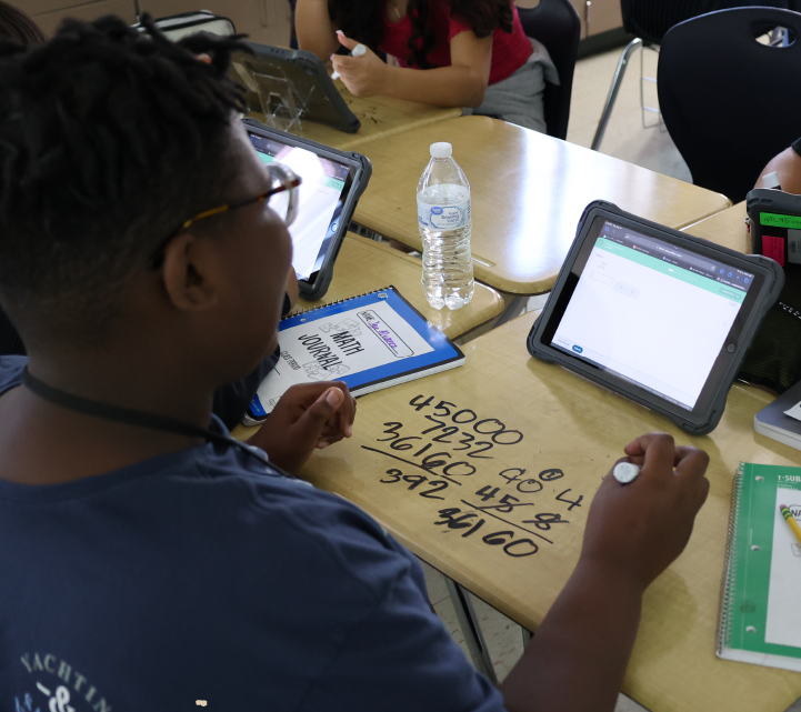 A young student is working on a math problem on a whiteboard, surrounded by various educational materials and electronic devices on the table.