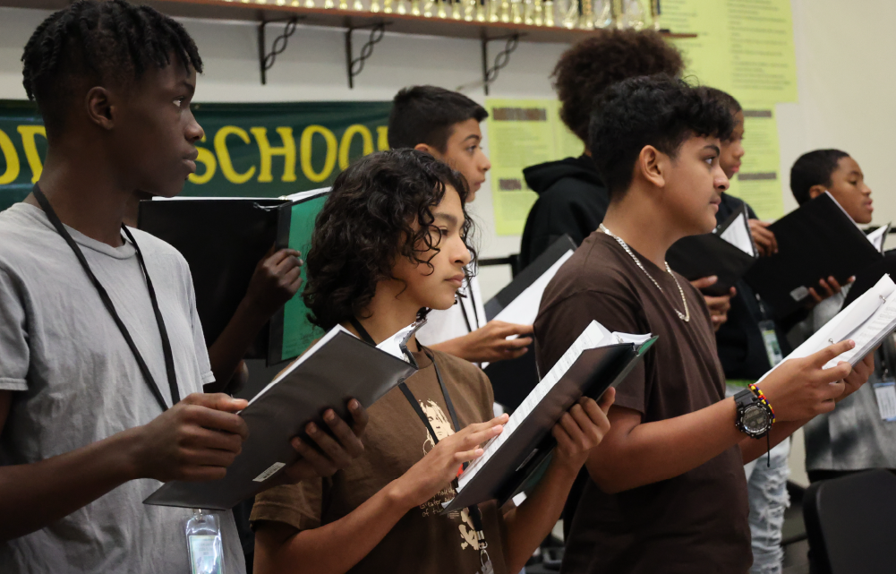 A group of young students, some with curly hair, standing together and holding books in a school setting with a "School" sign visible in the background.