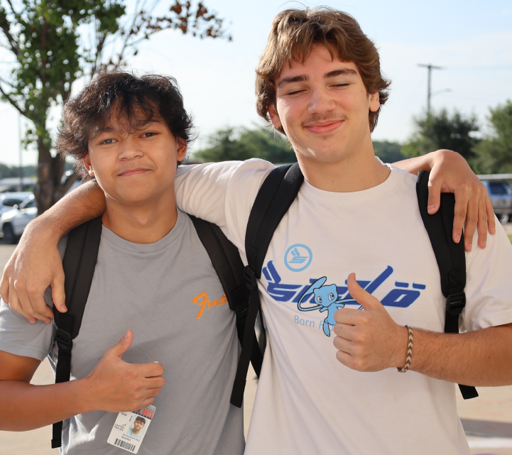 Two young men, one with curly hair and the other with straight hair, are standing together and smiling in an outdoor setting with trees and buildings visible in the background.