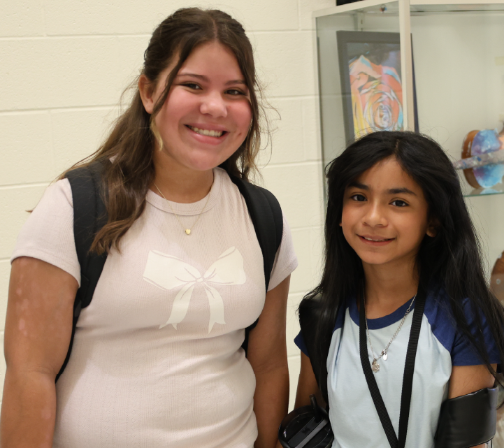 Two young women with friendly expressions are standing together in what appears to be a school or educational setting, with artwork and other decorations visible in the background.
