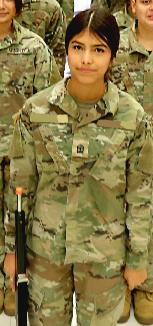 A person wearing military camouflage uniform stands in a setting with other military personnel and equipment in the background.