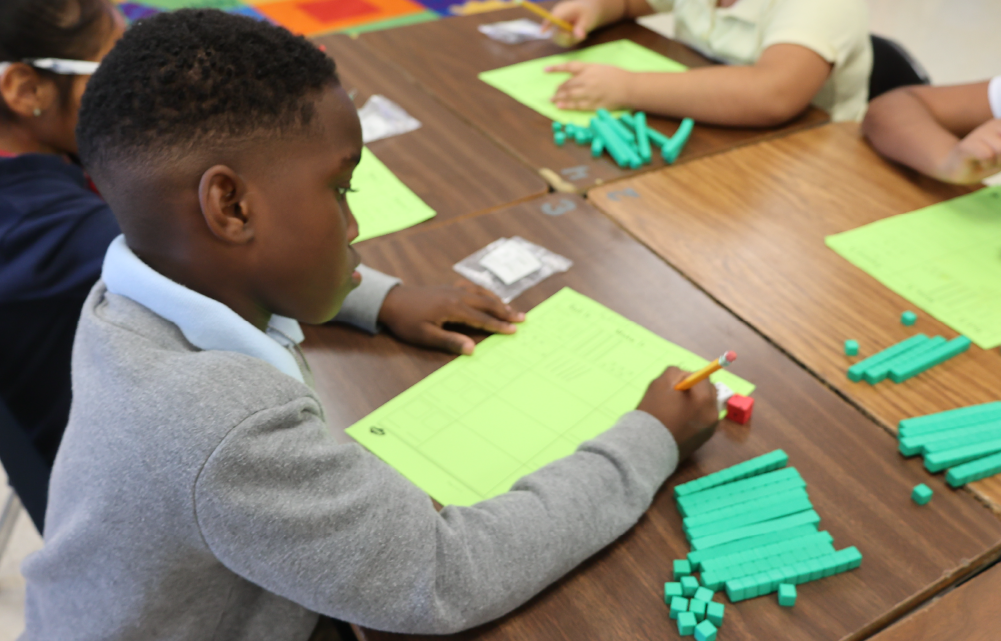 A young boy in a gray sweater is intently working on a craft project at a table with other children in the background.