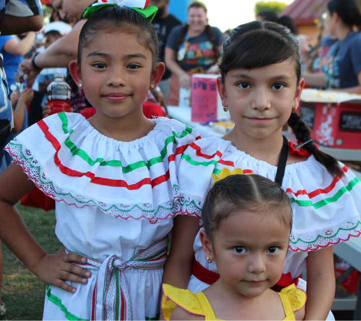 The image shows three young girls wearing traditional Mexican-style dresses, standing together in what appears to be an outdoor festival or celebration setting with people and tents visible in the background.