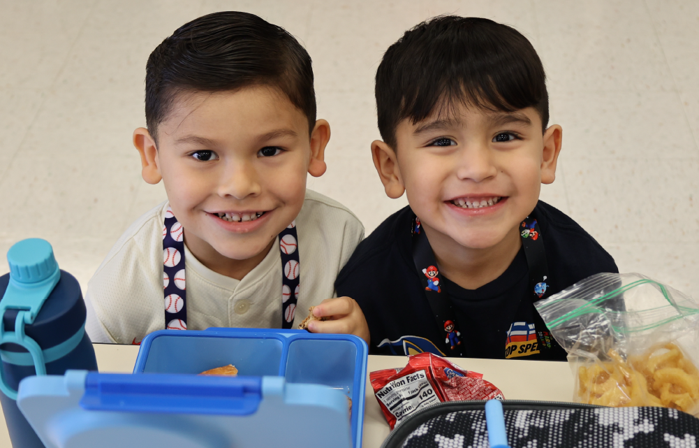 Two young boys with dark hair are sitting at a table, smiling and surrounded by various toys and snacks.