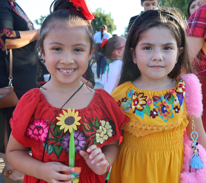 Two young girls wearing colorful, floral-patterned dresses are smiling and standing together in what appears to be an outdoor setting with other people in the background.