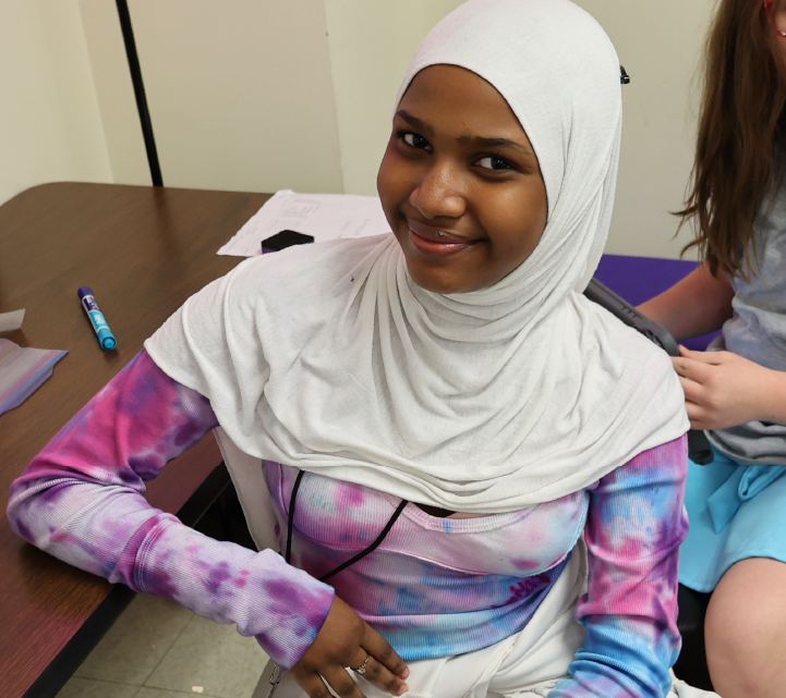A young woman wearing a white hijab and a colorful tie-dye shirt is sitting at a table, smiling at the camera.