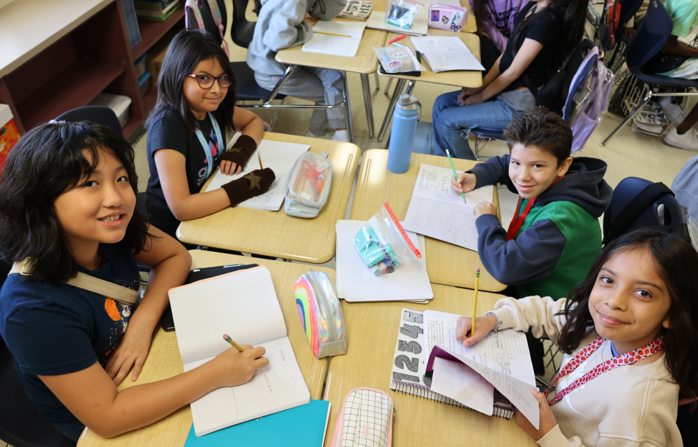 A group of young students sitting at desks, engaged in an art or drawing activity, with various art supplies and materials visible in the background.