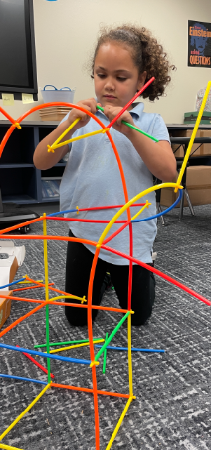 A young child is sitting on the floor, surrounded by a colorful array of plastic rods and connectors, seemingly engaged in a creative building activity in a classroom setting.