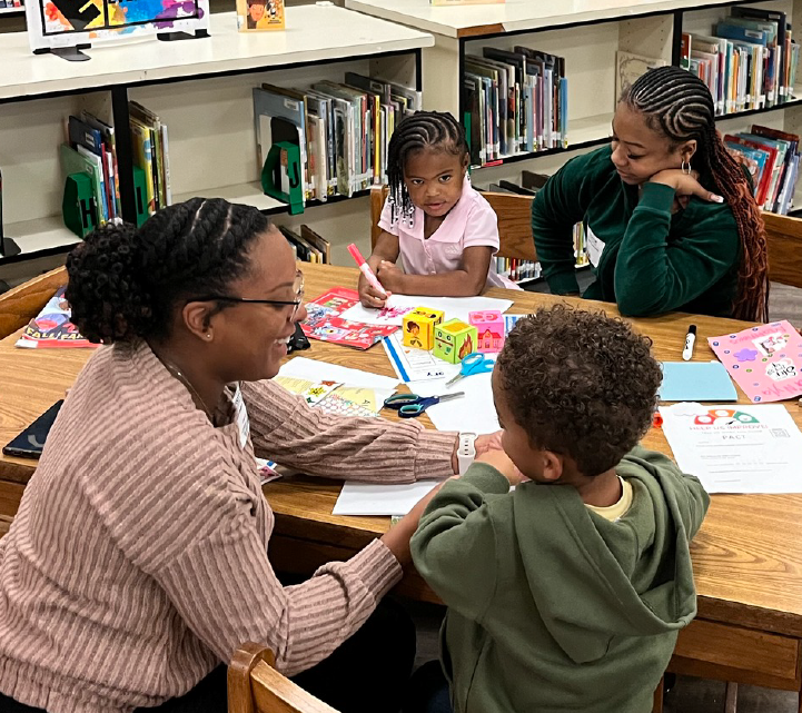 A group of young students are engaged in a learning activity at a table in a classroom setting, surrounded by bookshelves and educational materials.