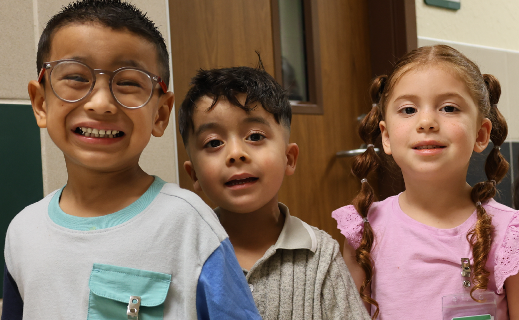 Three children, a boy wearing glasses and two girls, are smiling and posing together in what appears to be a classroom or school setting, with a chalkboard visible in the background.