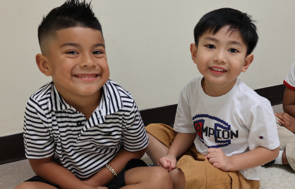 Two young boys, one wearing a striped shirt and the other a white t-shirt, are sitting together and smiling at the camera.