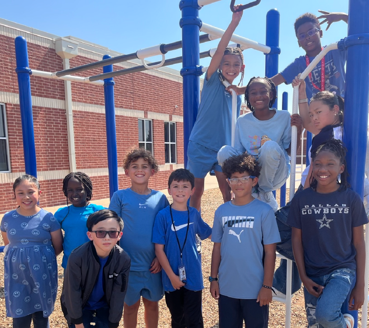 A group of children, both boys and girls, are gathered on a playground with a brick building in the background.