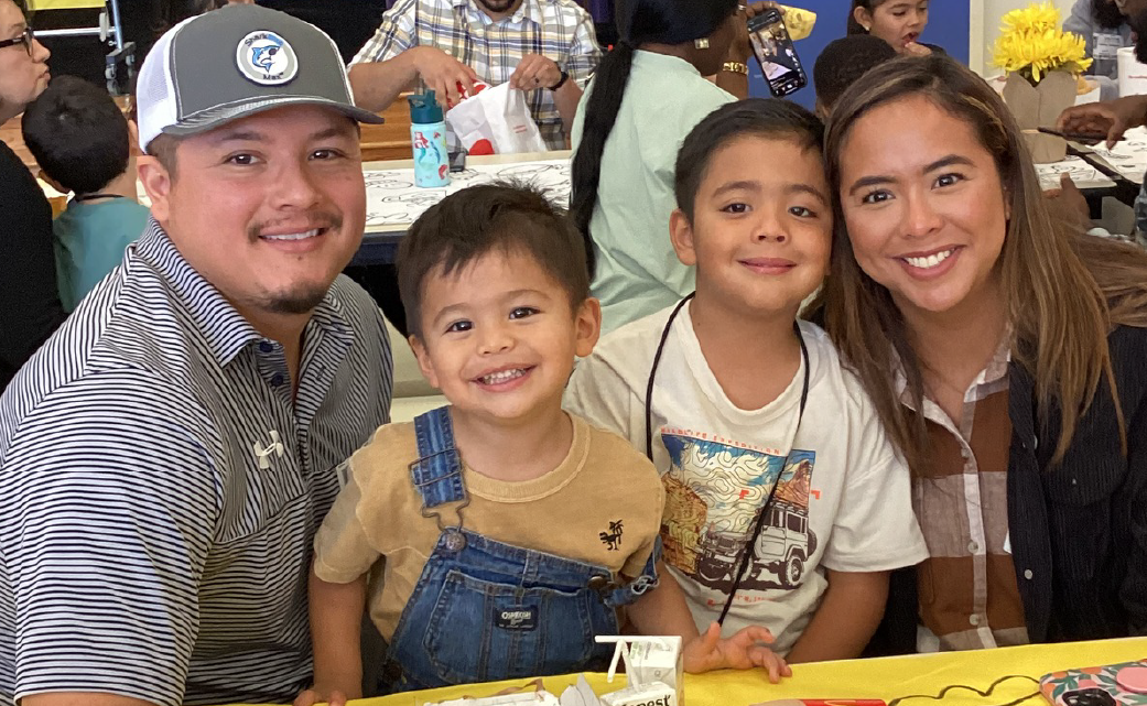 A happy family of four, including a man, a woman, and two young children, are smiling and posing for a photograph in what appears to be a casual setting.