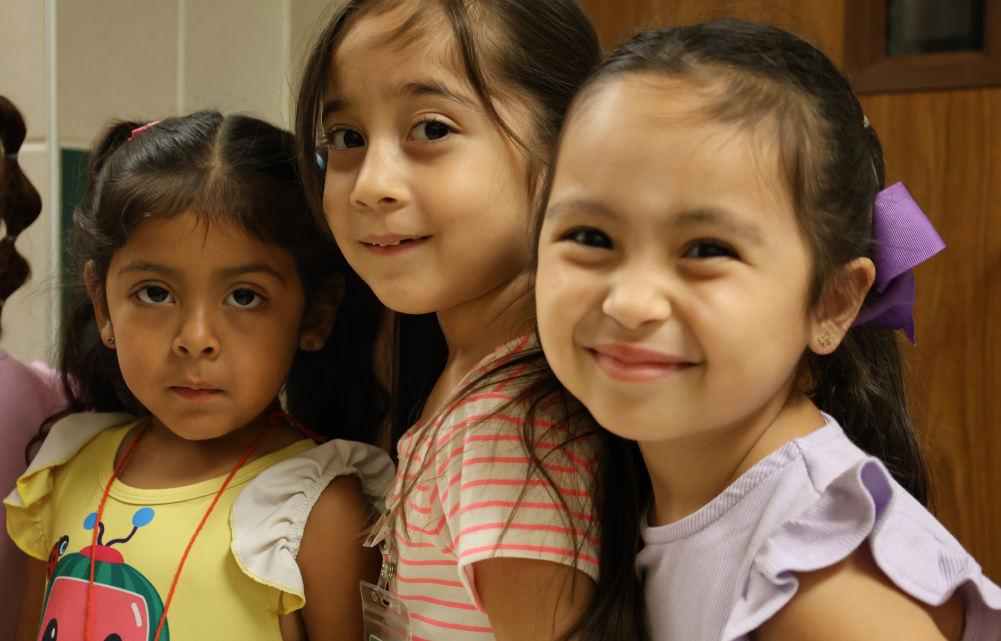 Three young girls with bright smiles and cheerful expressions, standing together in what appears to be a classroom or educational setting.