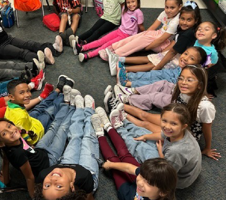 A group of young children, both boys and girls, are sitting together on the floor in what appears to be a classroom or daycare setting, with various toys and educational materials visible in the background.
