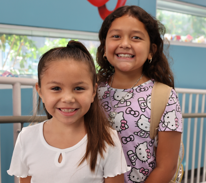 Two young girls with long dark hair are smiling and standing together in front of a blue wall with a window in the background.