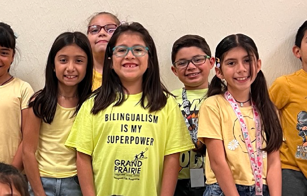 A group of young people, mostly women, wearing yellow shirts with the text "Bilingualism is my superpower" and "Grand Prairie" printed on them, standing together and smiling at the camera.