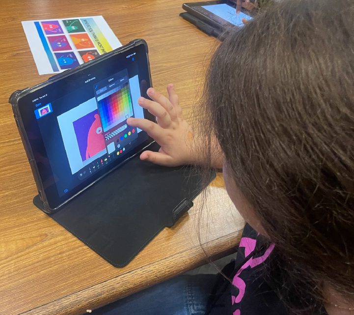 A person's hand is interacting with a tablet device displaying colorful shapes and patterns on a wooden desk.