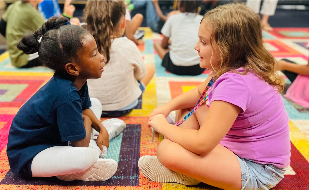 Two young girls, one with dark skin and the other with light skin, are sitting on the floor and engaging in conversation. The background appears to be a colorful, patterned rug or mat, suggesting an indoor setting.
