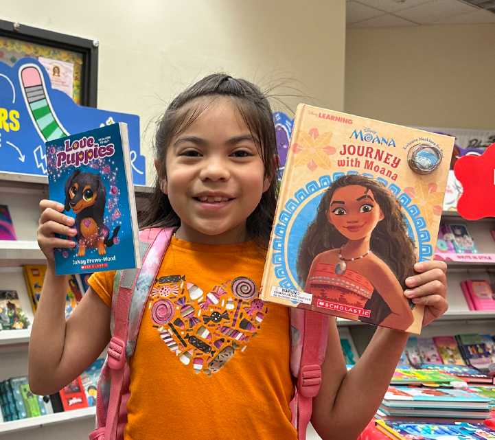 A young girl with a bright smile is holding two children's books, one featuring a character with dark skin and the other a character with light skin, in what appears to be a bookstore or library setting.