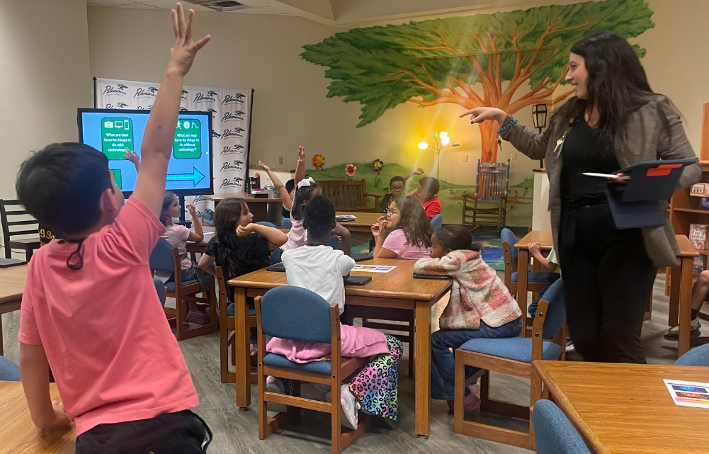 A classroom setting with students seated at desks, and a teacher standing in front of them, interacting with the class in an engaged manner.