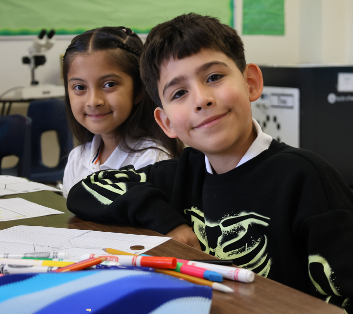 Two young students, a girl and a boy, are sitting at a desk in a classroom setting. They appear to be engaged in an activity or lesson, with various school supplies and materials visible on the desk in front of them.