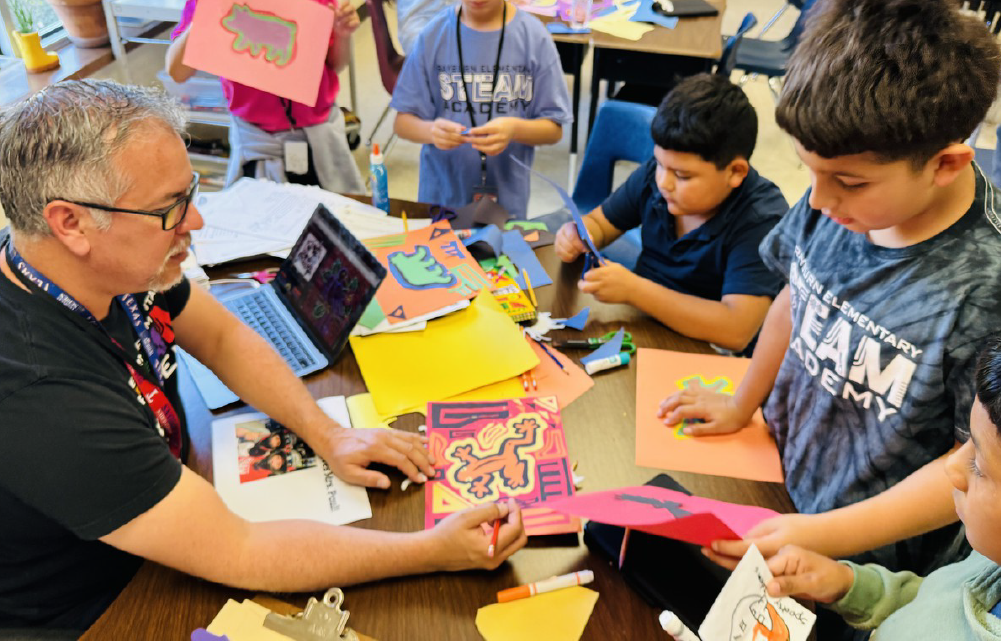 A group of students and a teacher are engaged in a collaborative arts and crafts activity, working together on a colorful project at a table in a classroom setting.