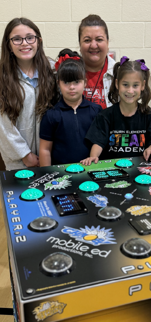 The image shows a group of people, including two adults and three children, gathered around a table with a large poster or display featuring various scientific or technological elements.
