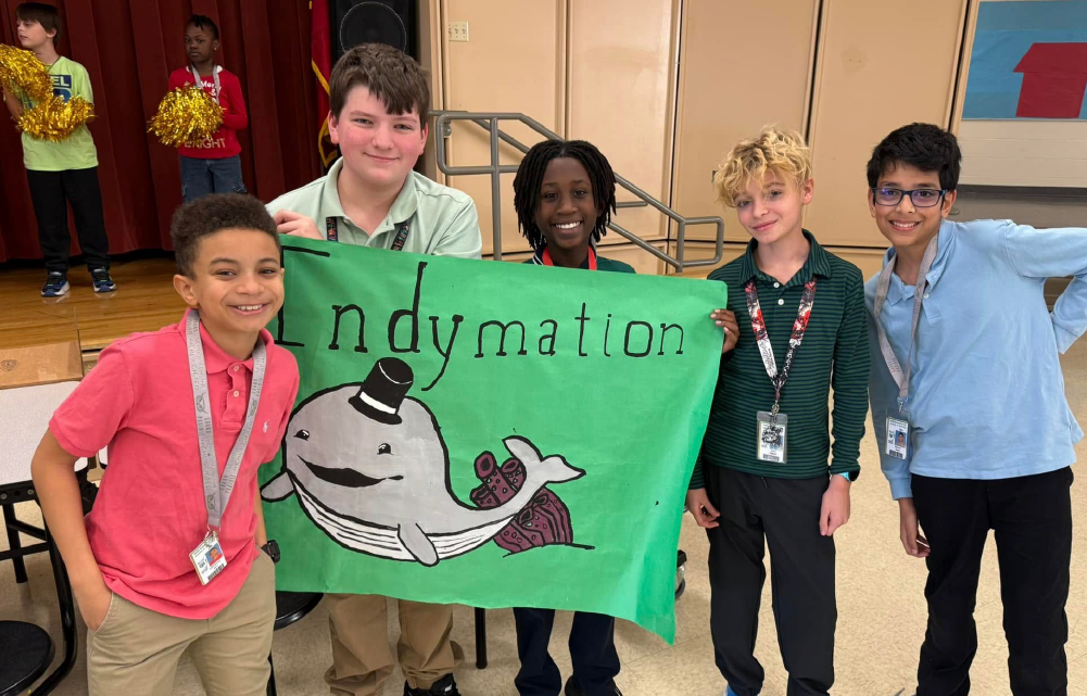 A group of diverse students, including both boys and girls, are holding a green banner with the word "Indymation" and an illustration of a whale.