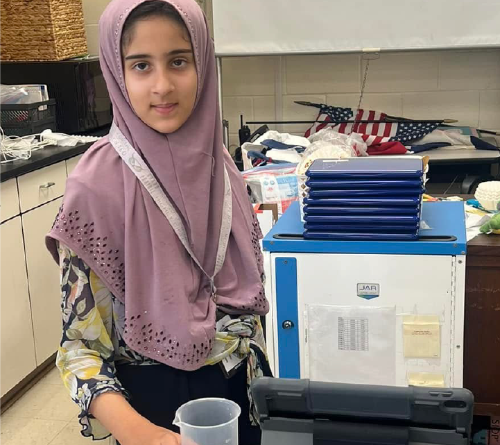 A young woman wearing a pink hijab stands in a room filled with various items, including a blue storage unit and an American flag.