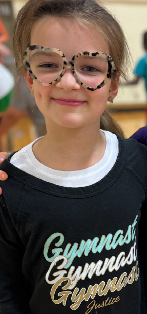 A young child wearing large, patterned glasses and a black t-shirt with the word "Gymnaast" printed on it, standing in what appears to be a classroom or indoor setting.