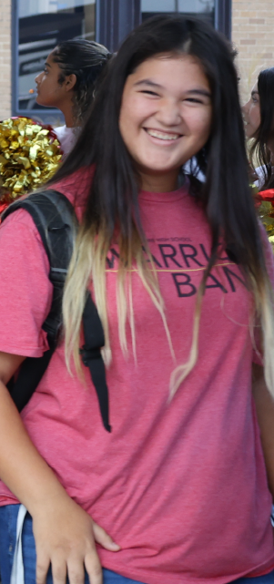 A smiling young woman wearing a pink "Marry Ban" t-shirt stands in the foreground, with a crowd of people visible in the background.