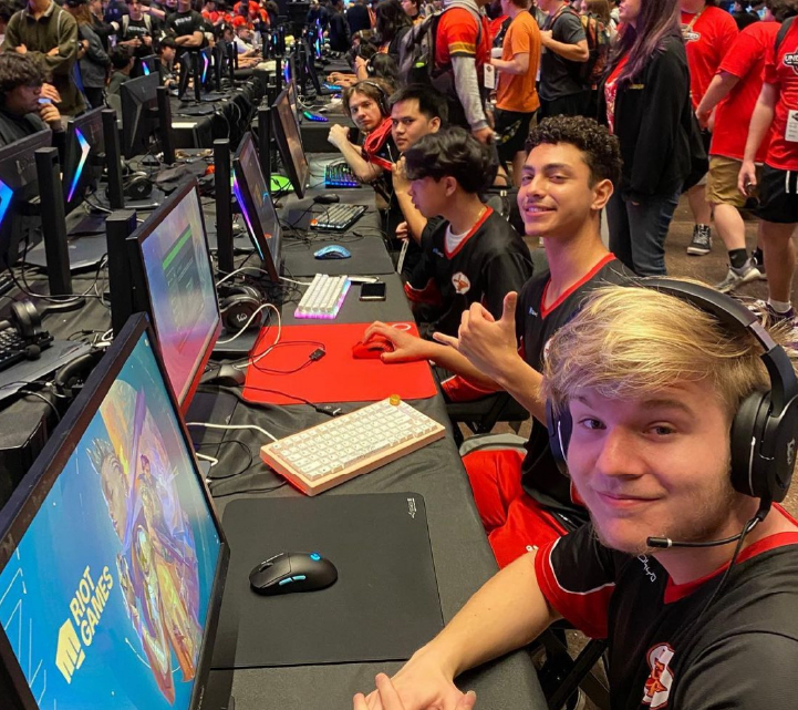 A crowded gaming event with people sitting at computers, some wearing headphones and engaged in gameplay, while others stand and observe in the background.