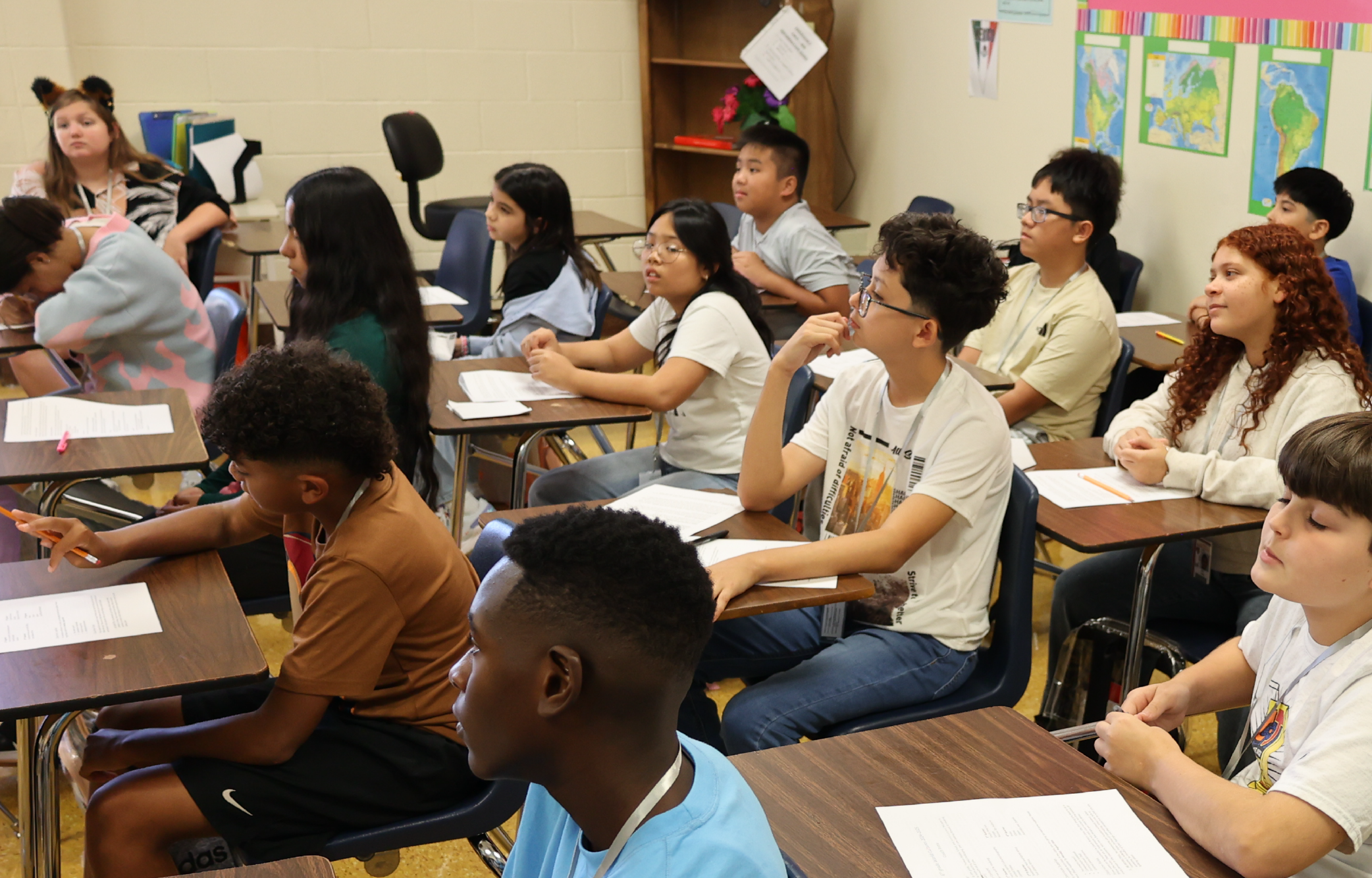 A classroom setting with students seated at desks, engaged in various activities, and a teacher standing at the front of the room.
