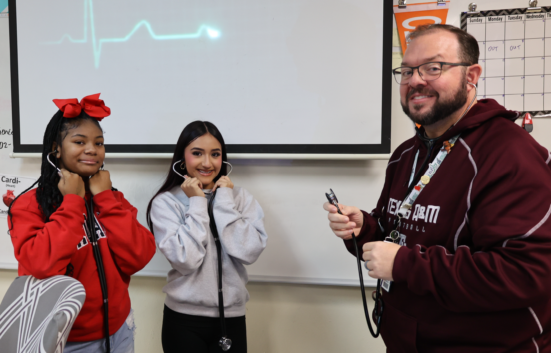 The image shows two young women wearing hoodies and a man with a beard standing in front of a projection screen.