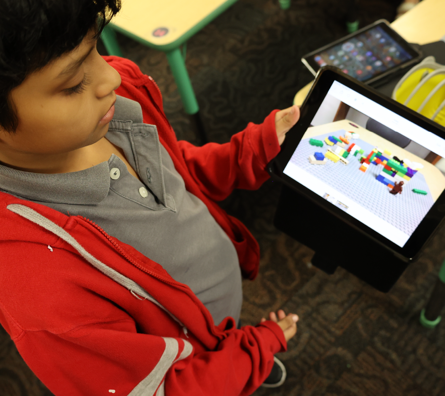 A young child in a red jacket is intently focused on a tablet device, surrounded by various toys and objects in the background.