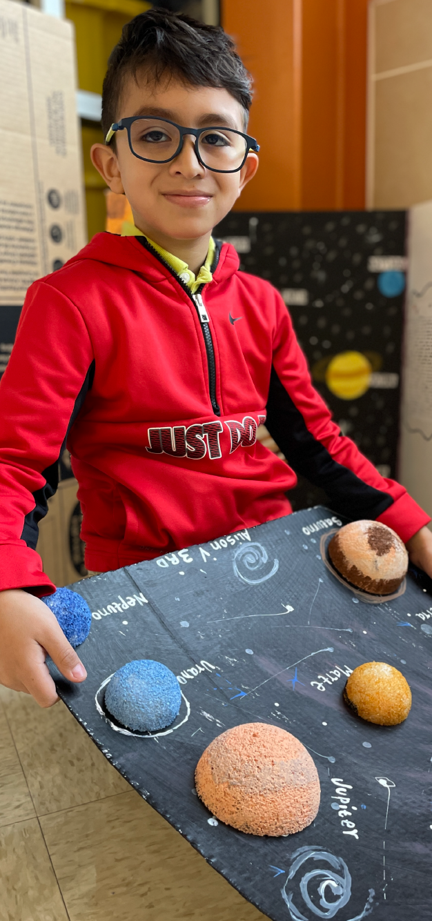 A young boy wearing a red shirt with the word "USTED" printed on it is sitting at a table, surrounded by various baked goods and a chalkboard-like surface with drawings of planets or celestial objects.