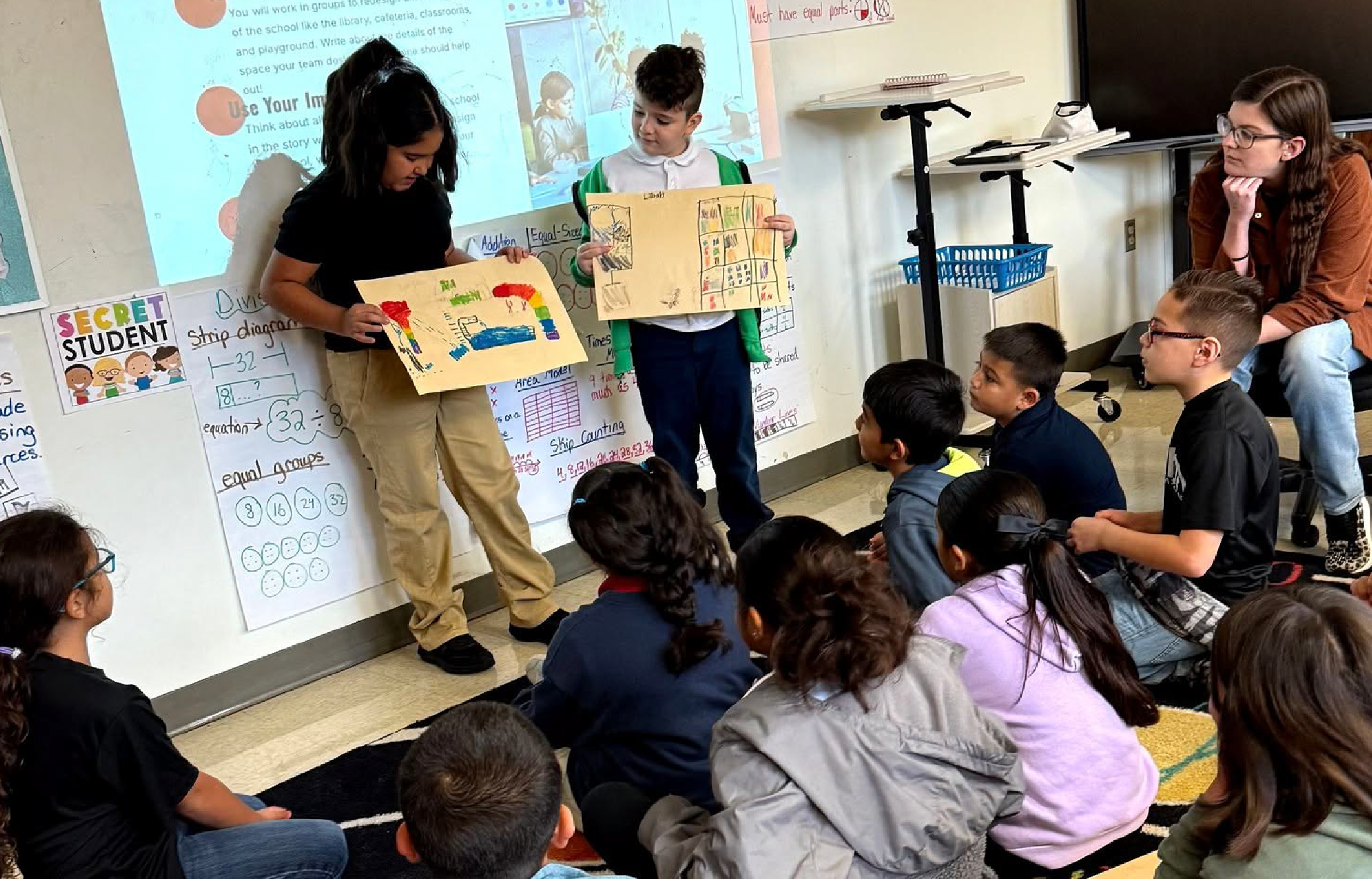 A group of students gathered in a classroom, with a teacher presenting a project or assignment to the class.