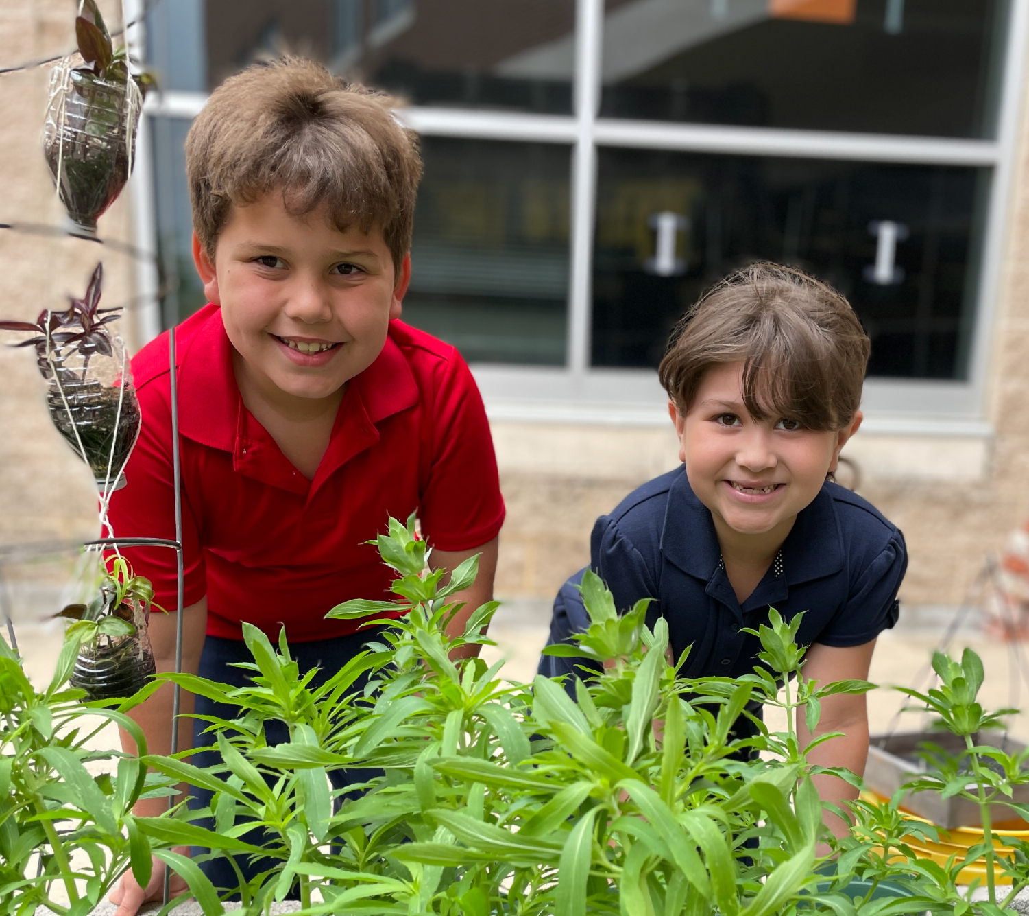 Two young boys, one wearing a red shirt and the other a blue shirt, are smiling and standing in front of a garden filled with lush green plants.